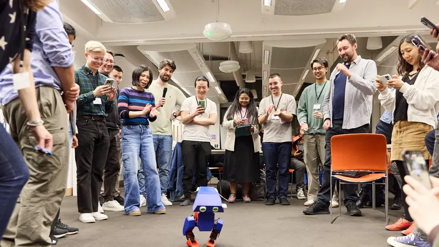 A large group of people standing in a semi circle taking photos and videos of a small blue robot 